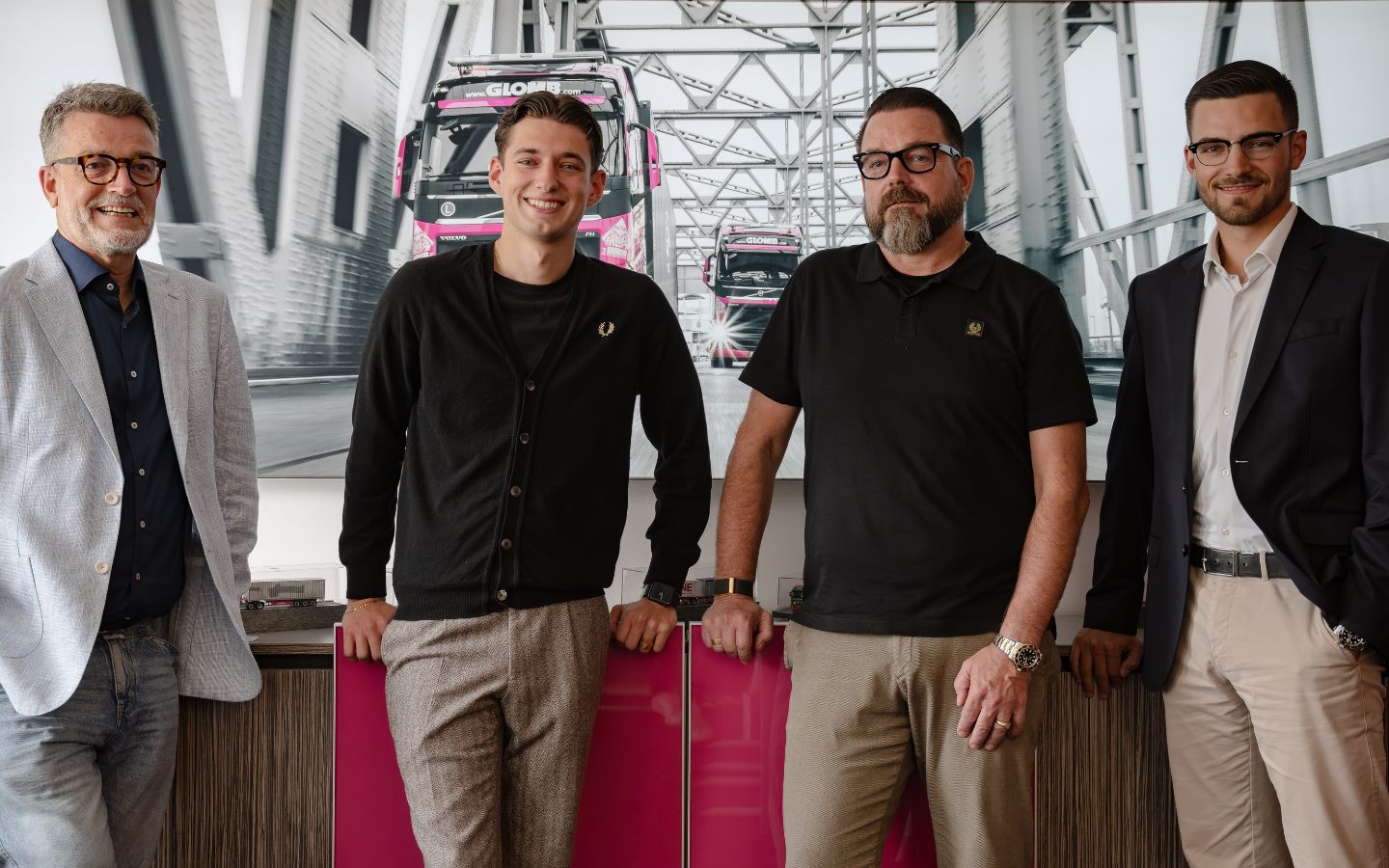 Vier Mitglieder der GLOMB Geschäftsführung und des Managements vor einem großformatigen Motiv mit pinken LKWs.
